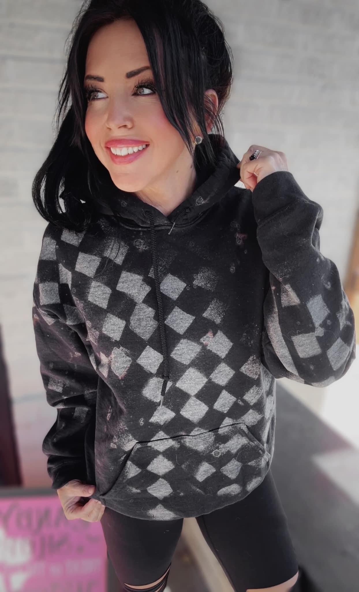 Black checkered hoodie with bleach splatter design, worn by a smiling woman with dark hair, styled for an edgy and casual streetwear look