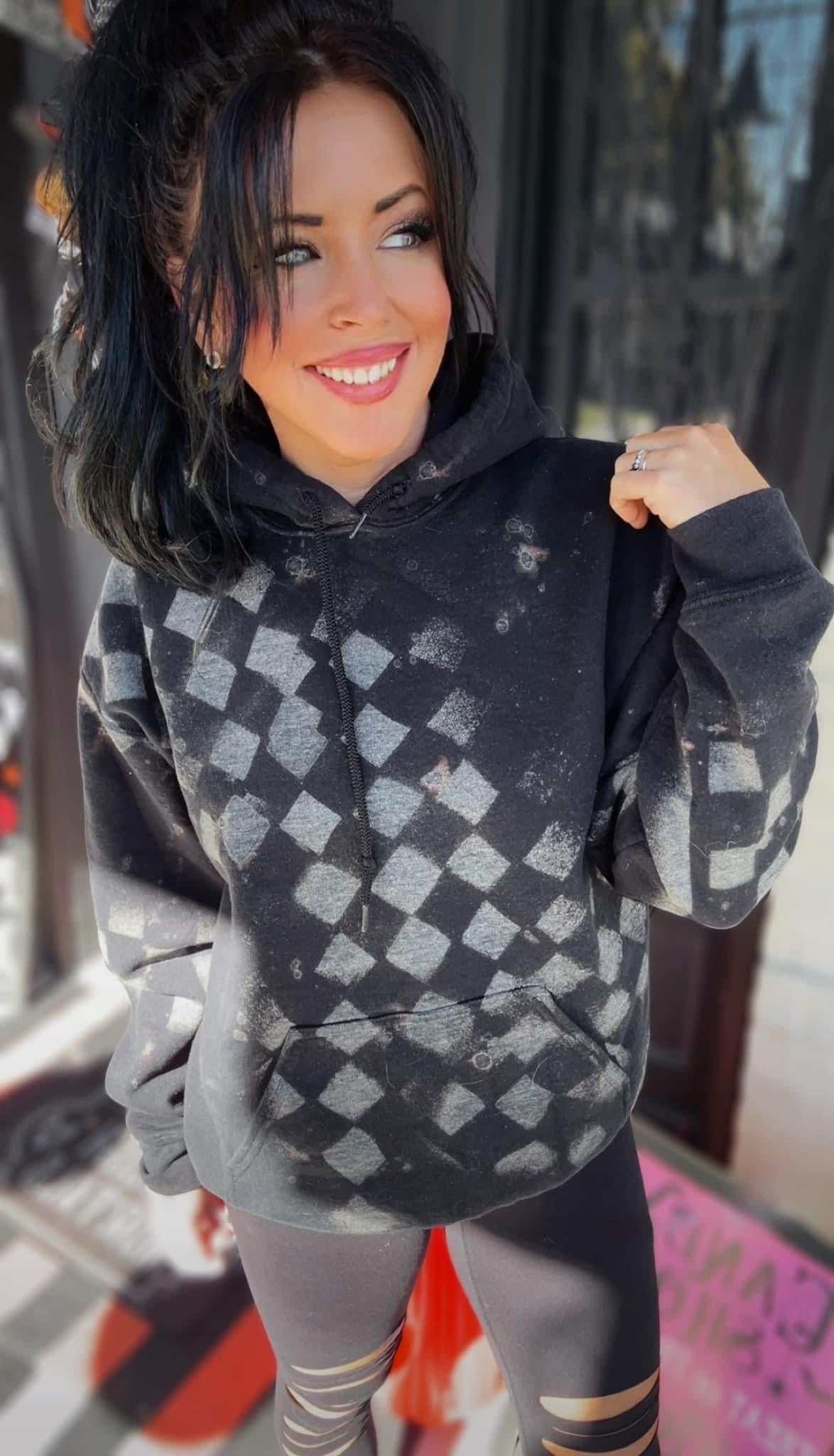 Black checkered hoodie with bleach splatter design, worn by a smiling woman with dark hair, styled for an edgy and casual streetwear look