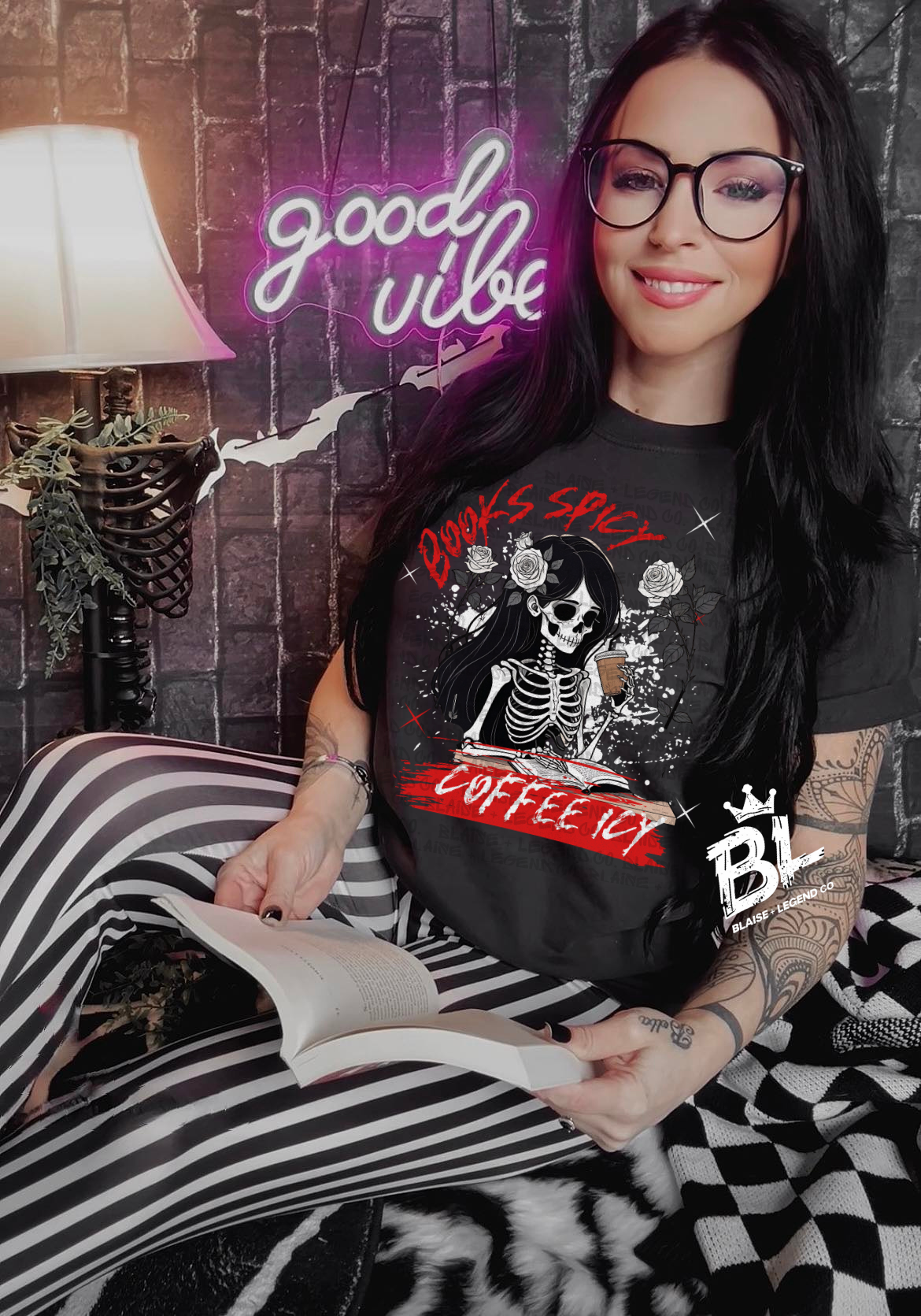 A woman wearing a black Comfort Colors t-shirt featuring a bold skeleton graphic surrounded by roses, an open book, and the text "Books, Spicy Coffee, and Vibes" in red and white. She is sitting in a cozy setting with striped leggings, holding a book, and smiling under a neon "Good Vibes" sign.