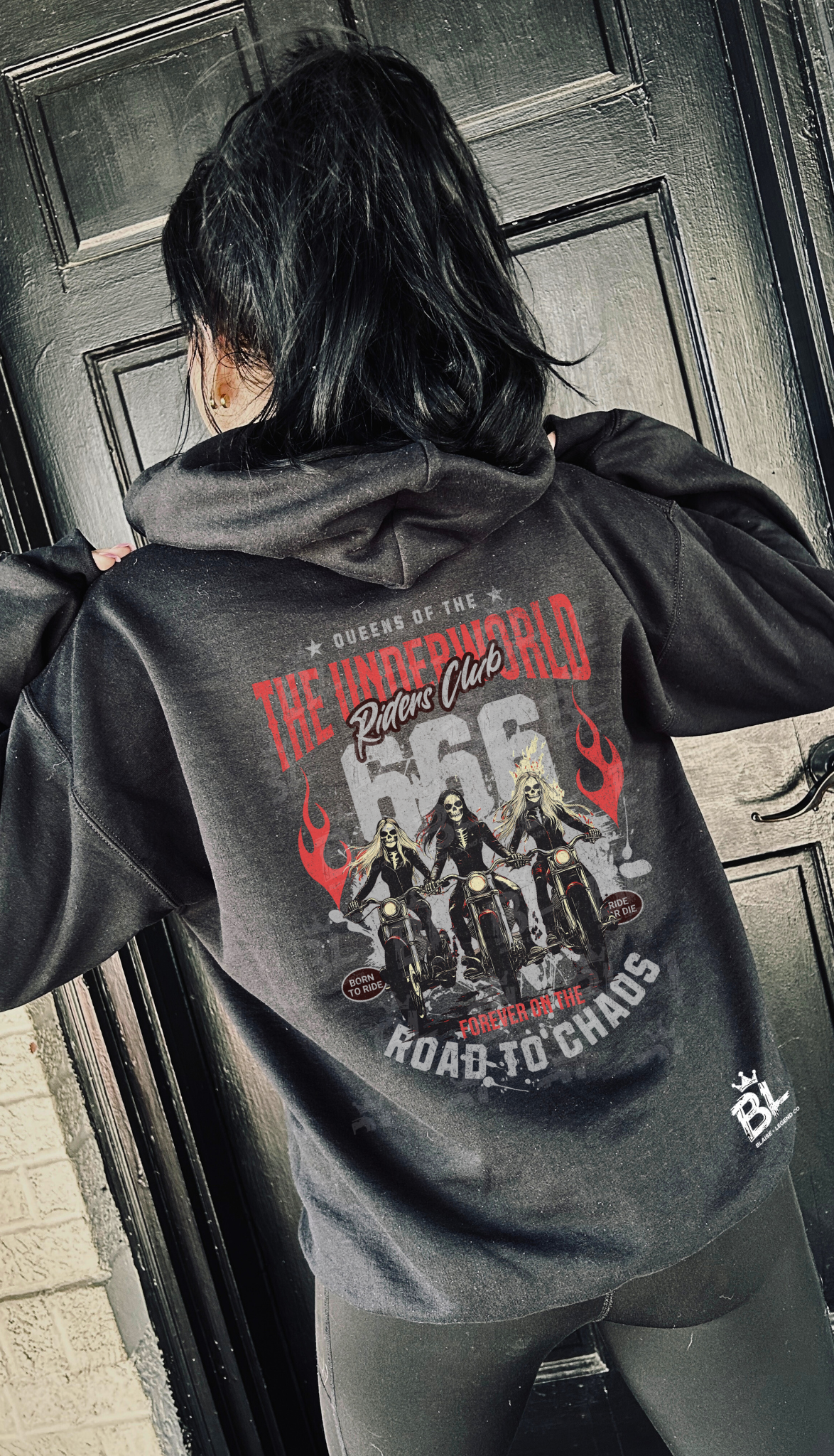 Back view of a woman wearing a black hoodie with 'Queens of the Underworld Riders Club' design featuring female bikers, red flames, and bold text, standing in front of a black door