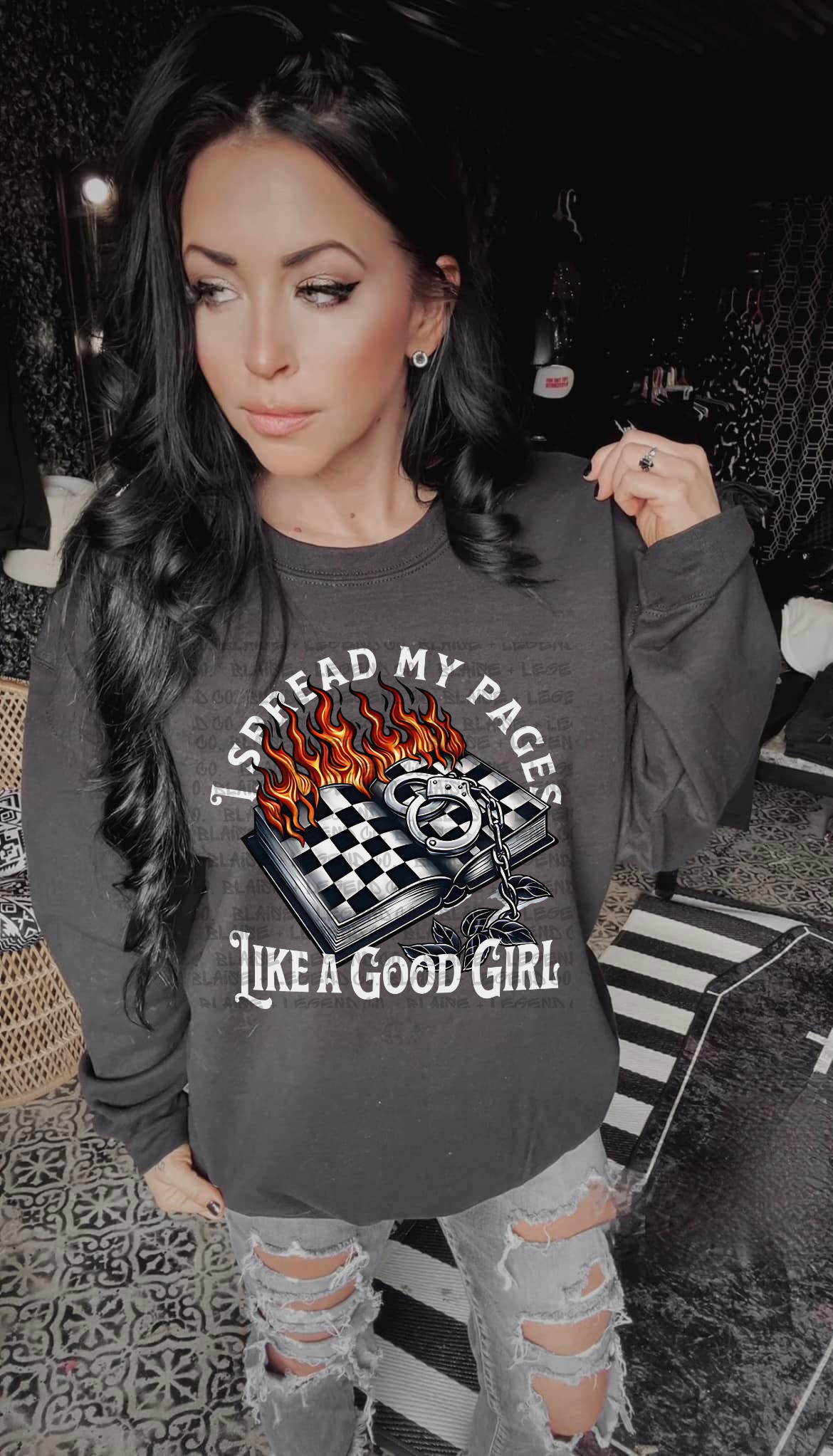 A woman with long black hair is wearing a charcoal grey crewneck sweatshirt featuring a bold design. The graphic shows an open book with flames, a checkered flag, handcuffs, and the text "I Spread My Pages Like a Good Girl." She stands in a stylish, edgy room with a dark aesthetic, pairing the sweatshirt with distressed jeans. This cozy and edgy sweatshirt is perfect for book lovers with a daring side.